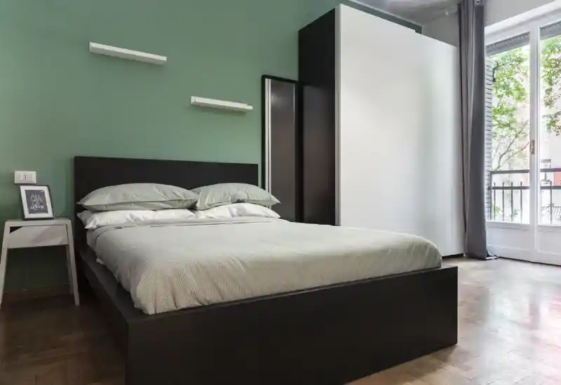 Well-lit, modern bedroom with a large double bed, simple bedside table and a wardrobe next to a balcony window. Neutral green accent wall and wood floor create a calm, stylish atmosphere.
