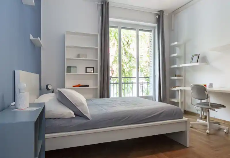 Bright, modern bedroom with a full-size bed, bedside table, shelving and a workspace next to a large window letting in natural light.