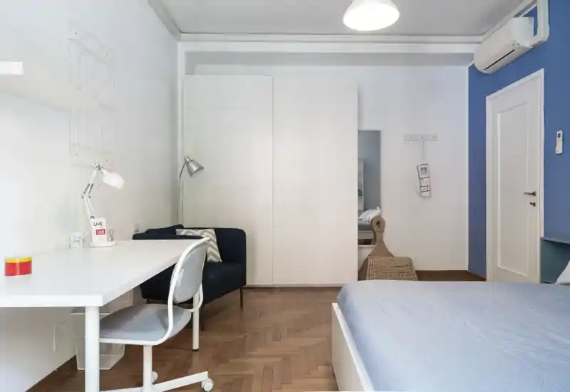 Bright, modern bedroom with a visible bed, white wardrobe, small desk and seating area — clean styling and warm parquet flooring.