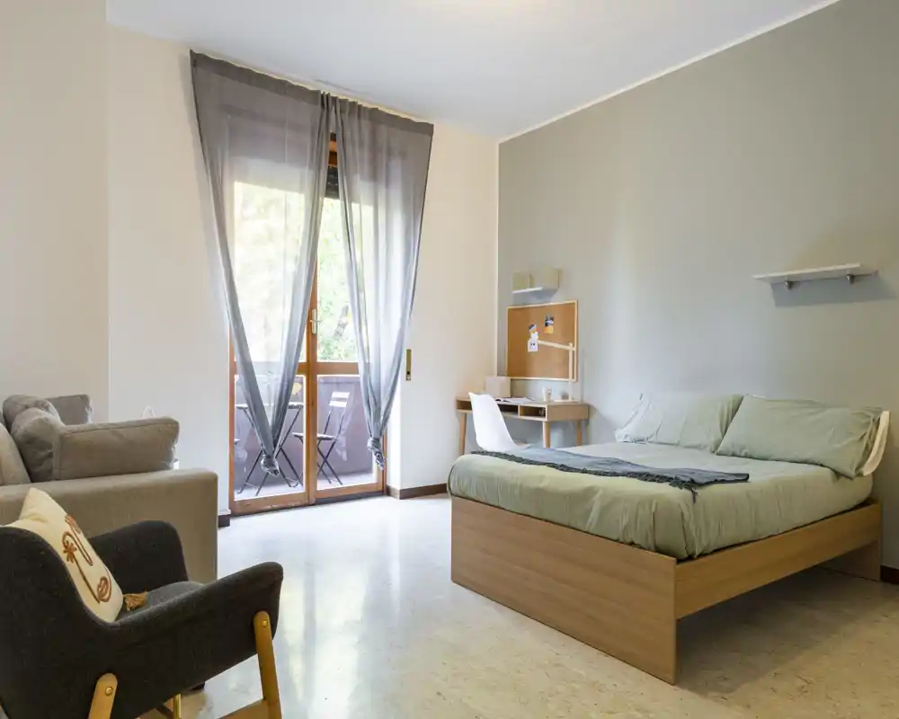 Spacious, bright bedroom with a double bed, a small work desk and seating area opening to a balcony — clean, modern lines and natural light make it ideal for comfortable stays.