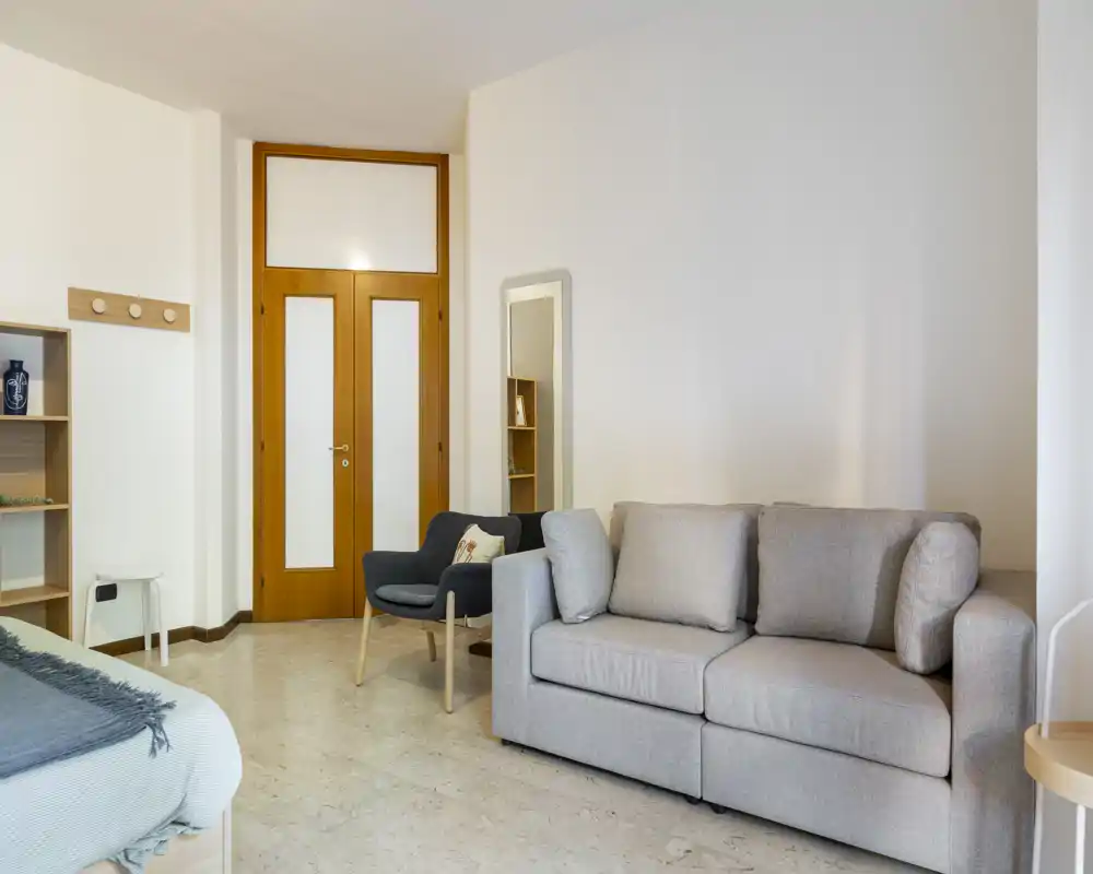Bright bedroom with a neutral-toned sofa and a partially visible bed in the corner, simple modern furnishings and soft textiles creating a calm, inviting space.