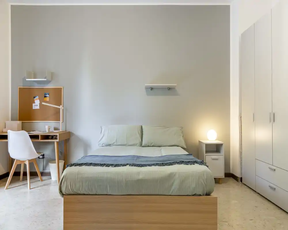 Bright, minimalist bedroom with a centrally placed double bed, bedside table with lamp and a small desk area — neat, calm and ready for guests.