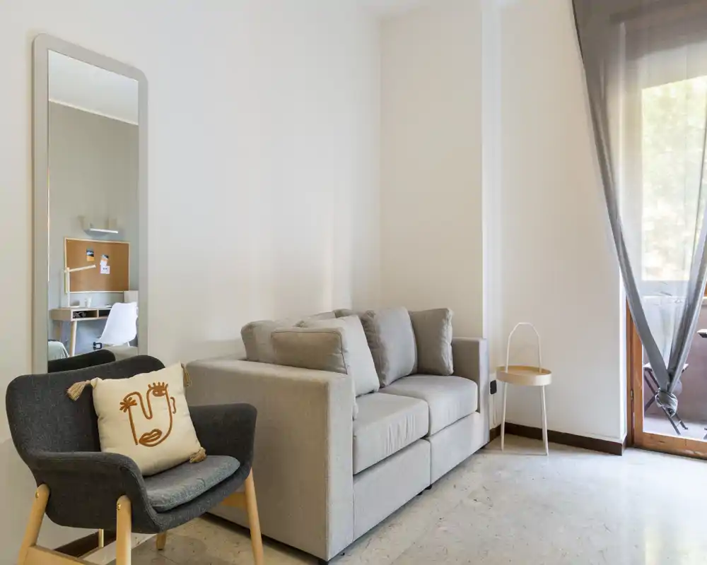 Bright, minimalist living area with a grey two-seater sofa, an armchair with decorative cushion and a full-length mirror; natural light enters from a balcony door.