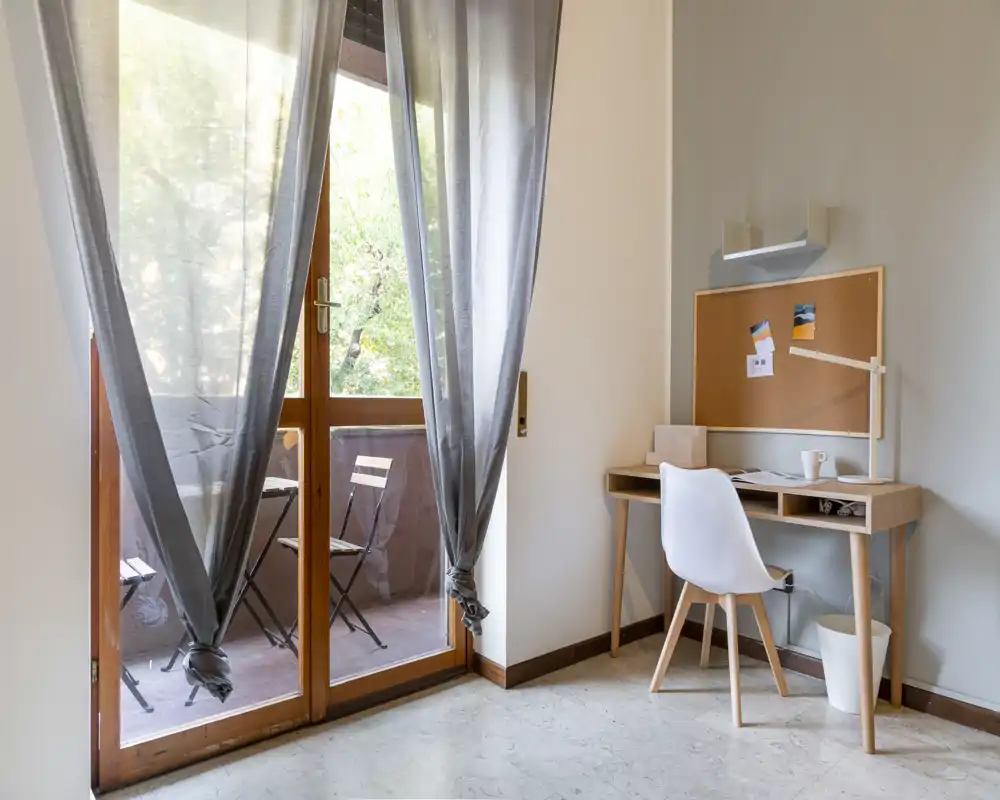 Bright, minimalist living area with a small wooden desk and chair beside large glass doors opening to a balcony — ideal as a light-filled home office corner.