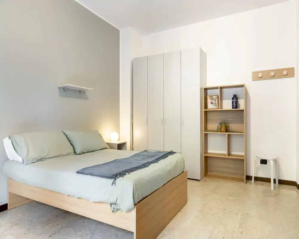Bright, minimalist bedroom with a wooden double bed, soft green linens, bedside lamp and spacious wardrobe — calm and welcoming sleeping space.