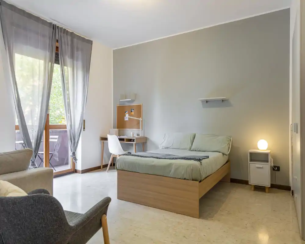 Bright, modern bedroom with a double bed, bedside table and a small work desk by the window that opens onto a balcony. Neutral tones and simple, tidy styling create a calm, inviting atmosphere ideal for a rental listing.
