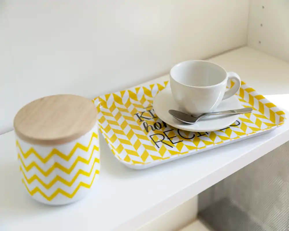 Primer plano de un rincón de cocina: una taza blanca con plato y cuchara sobre una bandeja amarilla estampada, junto a un tarro con tapa de madera.