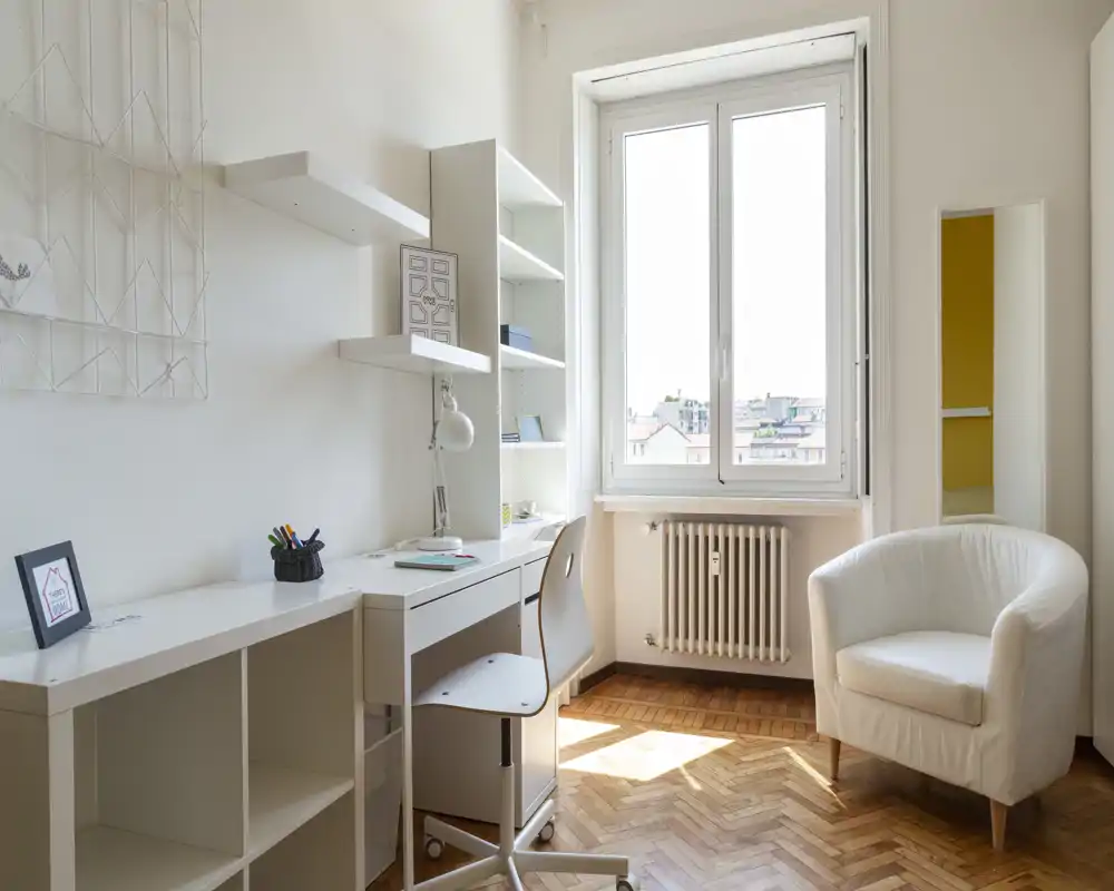 Zona de estar luminosa y minimalista con un sillón cómodo, escritorio y estanterías junto a una gran ventana que llena la habitación de luz natural — ideal para relajarse o trabajar.