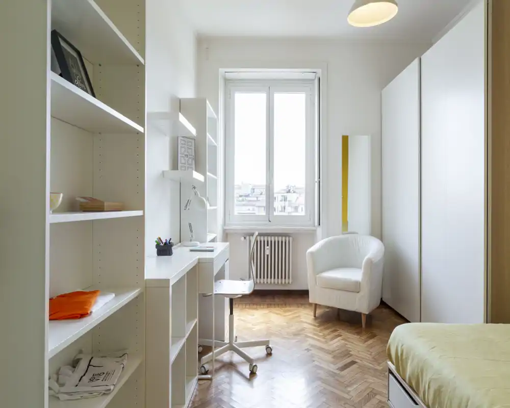 Dormitorio luminoso y ordenado con zona de estudio, butaca y gran armario — bien iluminado por una ventana grande y con un estilo limpio y minimalista.