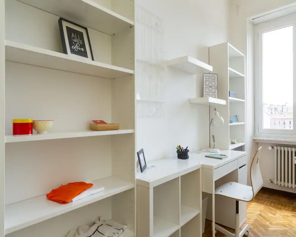 Espacio de trabajo moderno y luminoso con estanterías blancas, escritorio integrado y silla junto a una gran ventana que aporta luz natural.