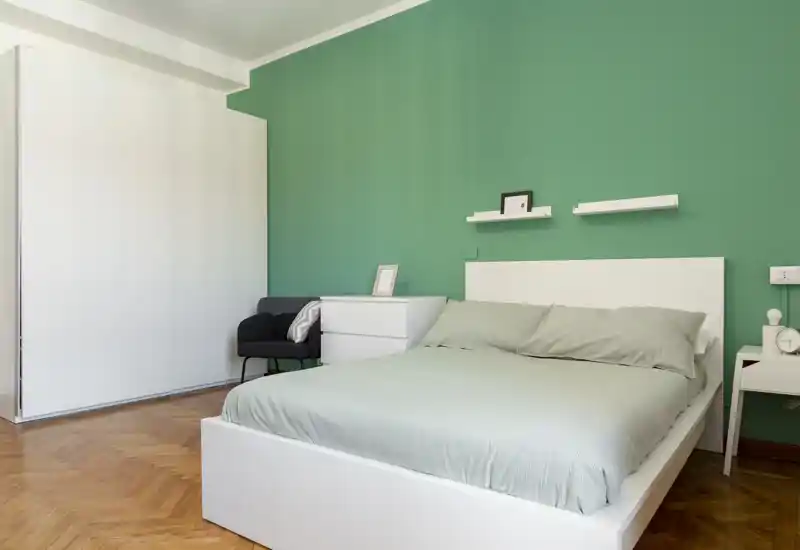 Camera da letto luminosa e minimal con letto matrimoniale davanti a una parete verde, mobili bianchi e pavimento in parquet — ambiente moderno e rilassante.