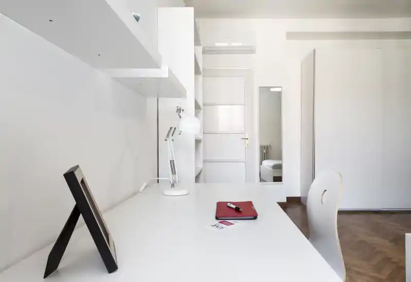 Spazio letto minimalista e luminoso con scrivania bianca, lampada e sedia; un armadio e la porta con specchio mostrano una piccola porzione del letto riflessa. Linee pulite e pavimento in parquet creano un ambiente moderno e tranquillo.