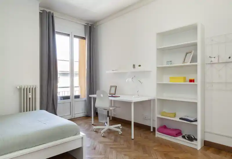 Camera da letto luminosa e minimal con un letto singolo, scrivania vicino alla finestra e una libreria alta. La luce naturale e il parquet pulito creano un'atmosfera moderna e tranquilla, perfetta per affitti brevi.