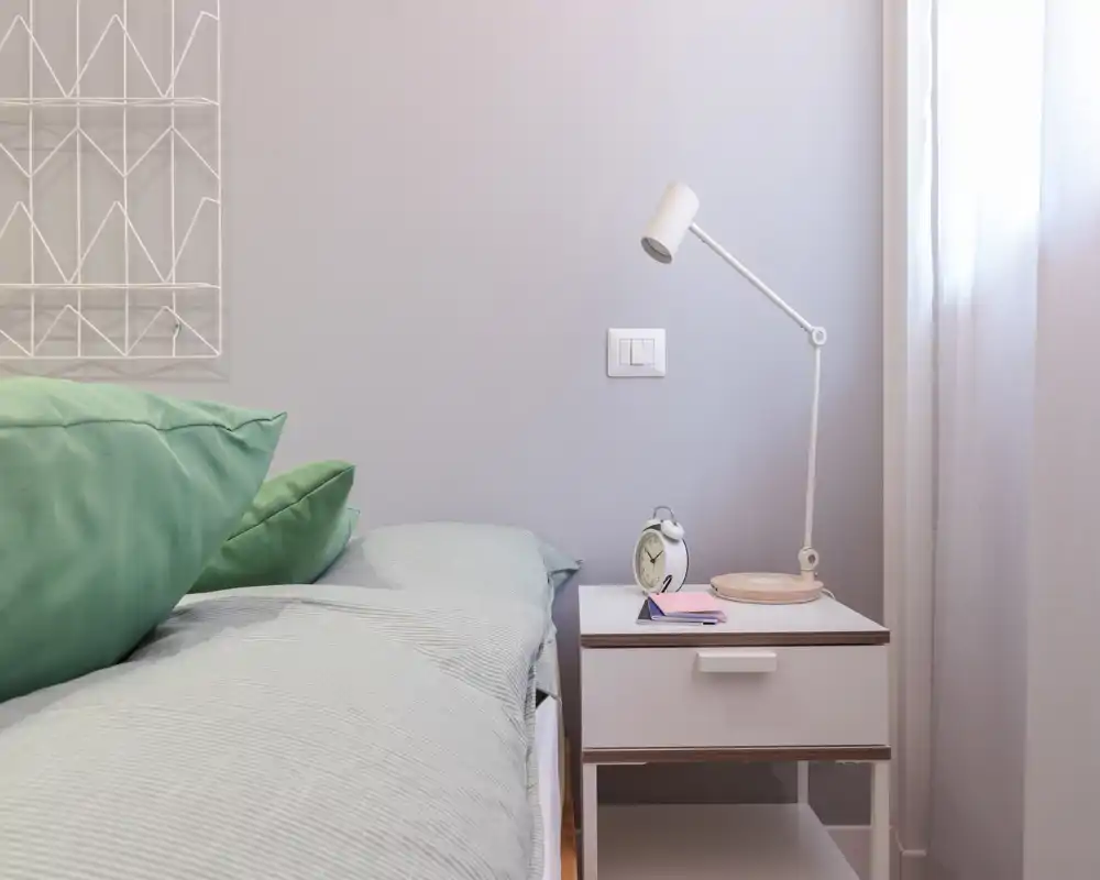 Angolo camera luminoso e tranquillo con letto ben rifatto, cuscini verdi come dettaglio e un comodino minimal con lampada e sveglia — ideale per un soggiorno riposante.