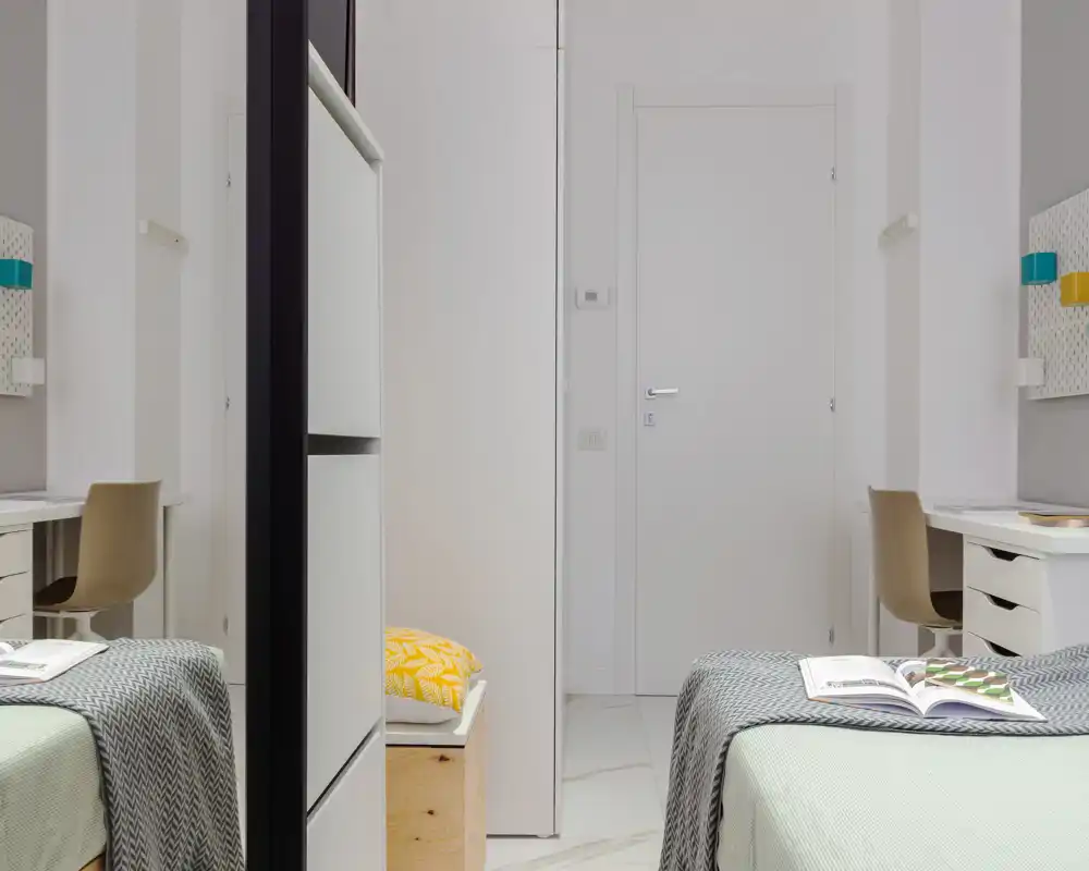 Camera con due letti singoli, biancheria ordinata, contenitori e scrivanie su entrambi i lati; stile luminoso e minimale con buona simmetria, ideale per affitti condivisi.