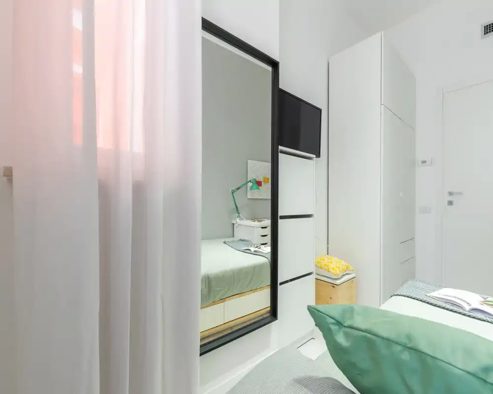 Camera da letto luminosa e moderna con letto ben rifatto, tessili morbidi e un grande specchio a parete che amplia lo spazio; mobili bianchi e accenti verde chiaro per un’atmosfera fresca e rilassante.