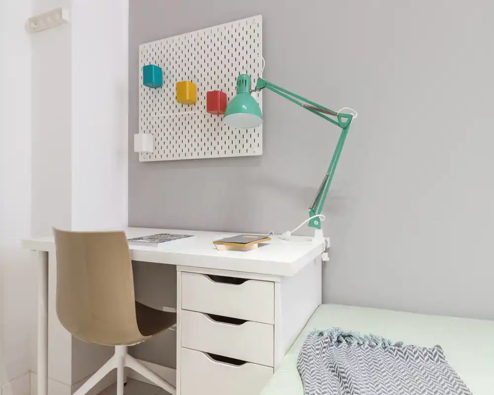 Angolo di camera moderno con scrivania bianca, lampada da lavoro e una porzione del letto visibile. Spazio luminoso e pulito con dettagli colorati minimal.
