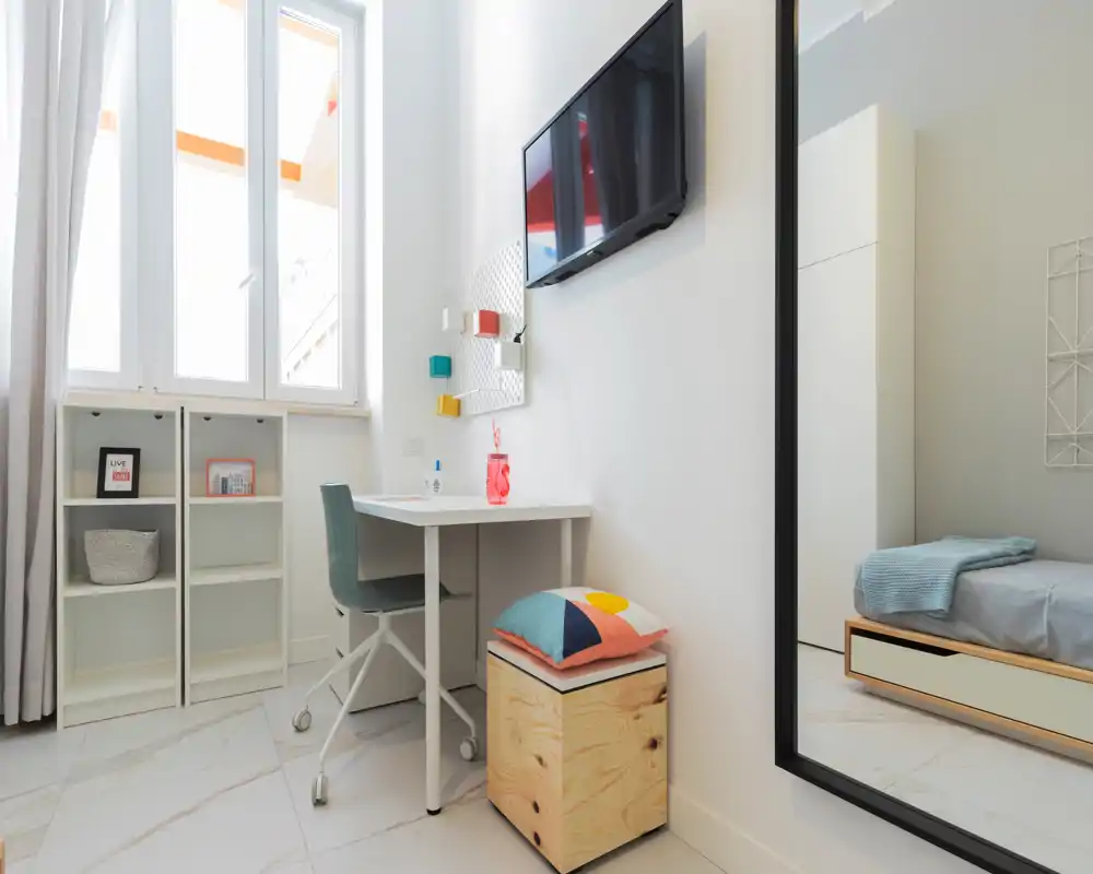 Camera da letto moderna e luminosa con una scrivania minimal, scaffali e il letto parzialmente visibile nello specchio; finiture bianche e cuscini colorati creano un tocco fresco.