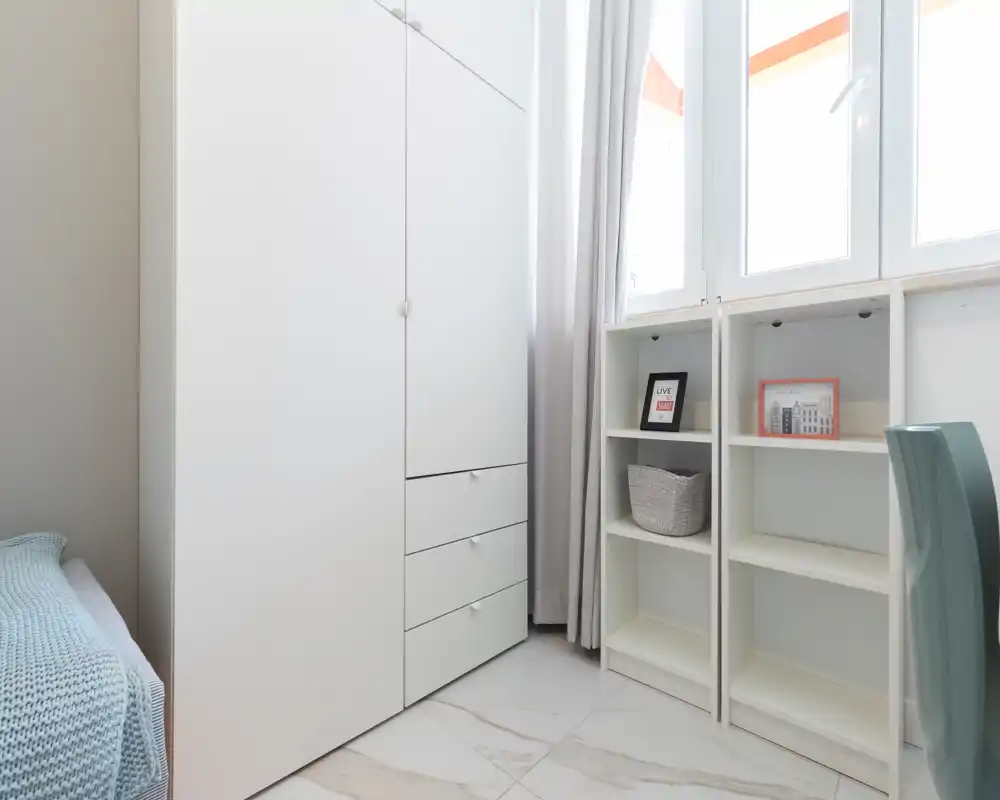 Angolo di camera da letto compatto e luminoso con un armadio bianco, una scaffalatura e il letto parzialmente visibile a sinistra, con una coperta azzurra e ampie finestre che forniscono luce naturale.