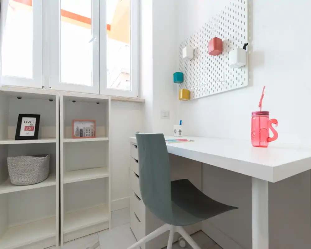 Angolo studio luminoso e minimal con scrivania bianca, sedia ergonomica, scaffali e pannello a parete con contenitori colorati — ideale per lavoro da remoto o studio.