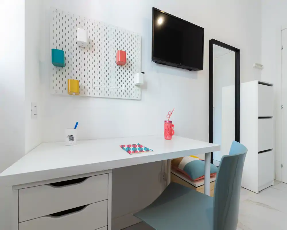Angolo studio moderno e luminoso con scrivania bianca, sedia, TV a muro e contenitori decorativi; ideale per studio o smart working.
