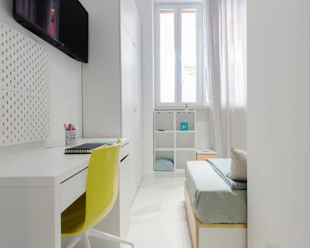 Camera singola compatta e moderna con scrivania integrata, finestra luminosa e armadi minimal; biancheria in toni pastello e sedia gialla per un tocco di colore.