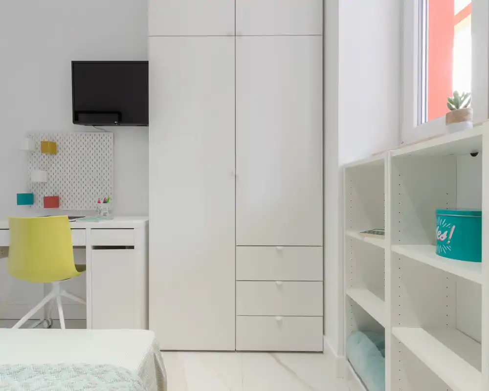 Angolo di camera da letto luminoso e moderno con armadio bianco, scaffalatura aperta e una piccola scrivania. Il letto è parzialmente visibile in primo piano e la stanza ha linee pulite e luce naturale.