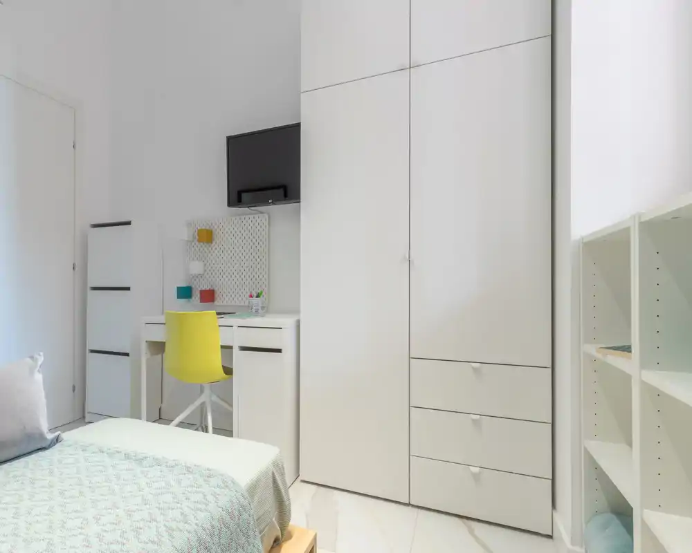 Camera da letto luminosa e minimal con letto singolo visibile, armadio a muro e una piccola scrivania con sedia gialla — perfetta per una persona o uno studente.