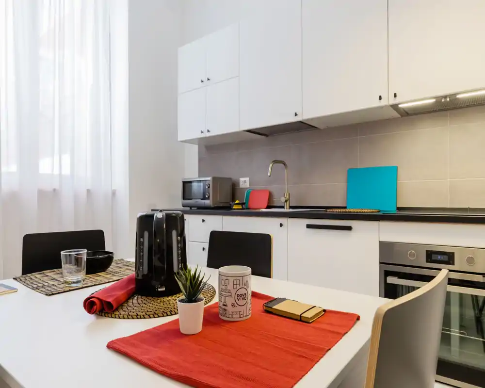 Cucina moderna e luminosa con pensili bianchi, forno incasso, lavello e tavolo da pranzo ben apparecchiato con dettagli rossi e una piccola pianta — perfetta per appartamenti compatti.