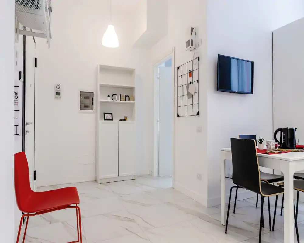 Zona giorno luminosa e moderna con pareti bianche minimal, piccolo tavolo da pranzo con sedie, TV a parete e decorazioni semplici — perfetta per soggiorni in città.