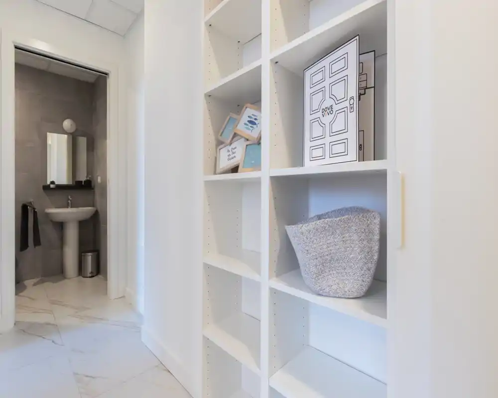 Ingresso/corridoio luminoso e moderno con scaffalature bianche incassate e pavimento piastrellato, con vista su un bagno compatto con lavabo a colonna.
