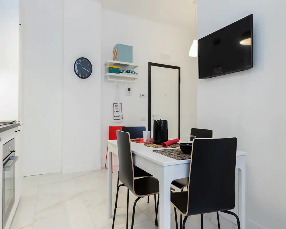 Zona pranzo moderna e luminosa con tavolo bianco per quattro, sedie nere dal design contemporaneo, TV a muro e arredo minimalista.