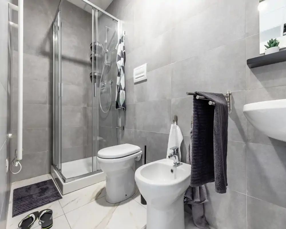 Bagno moderno e compatto con doccia in vetro, WC, bidet e lavabo a parete; piastrelle grigie neutre e sanitari bianchi per un aspetto pulito e contemporaneo.