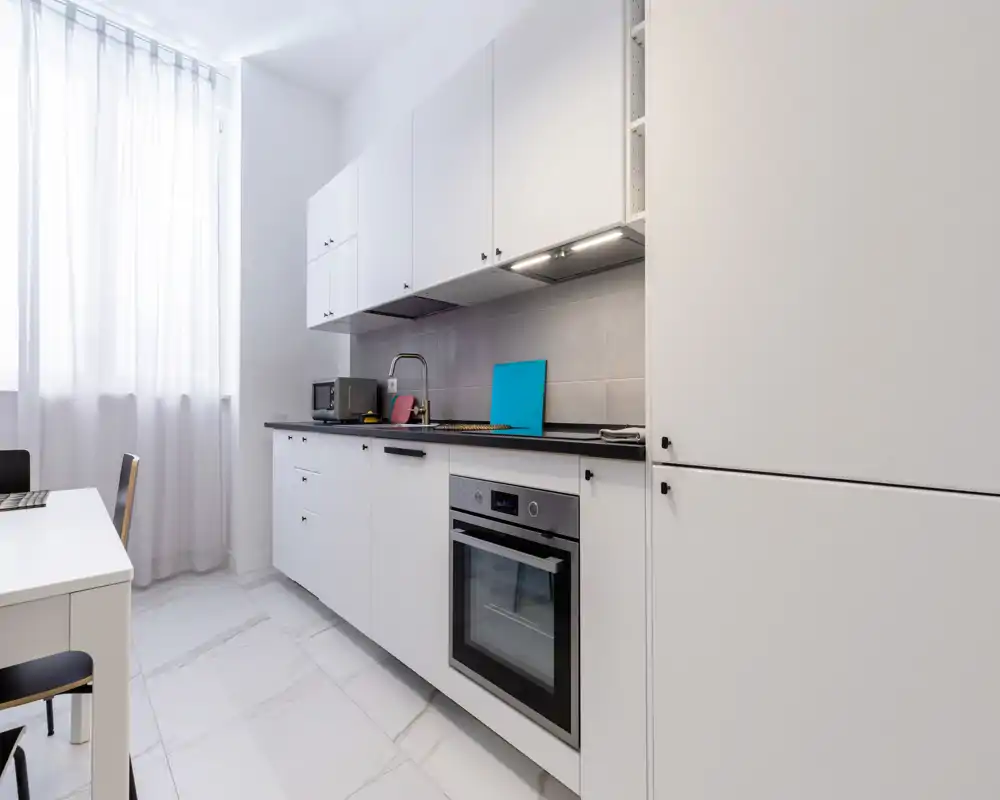 Cucina moderna e luminosa con mobili bianchi, forno integrato e piano lavoro nero pulito. La luce naturale da una grande finestra valorizza il design minimal e lo spazio ordinato.