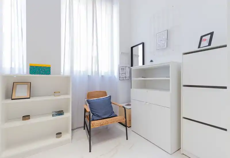 Bright, minimal living area with a wicker armchair and white storage units; sheer curtains let in soft natural light for a calm, airy atmosphere.