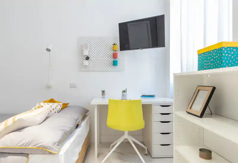 Angolo camera da letto moderno e luminoso con letto singolo ben rifatto, scrivania bianca e sedia gialla sotto la TV a parete — spazio compatto, pulito e ben illuminato.