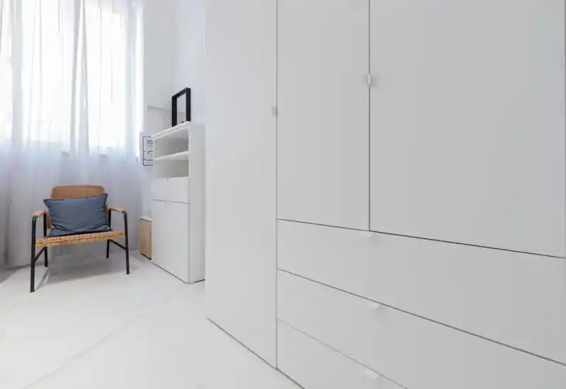 Angolo camera luminoso e minimale con grande armadio bianco, poltrona in vimini e tende leggere che lasciano entrare la luce naturale — pulito, moderno e arioso.