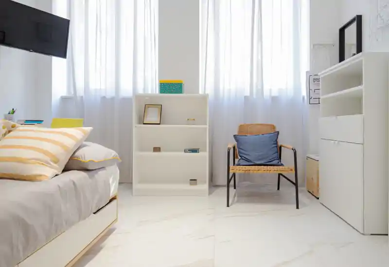 Camera da letto luminosa e minimalista con letto accogliente, cuscini morbidi e luce naturale filtrata dalle tende leggere; scaffalatura funzionale e poltrona in rattan che aggiungono calore e spazio contenitivo.