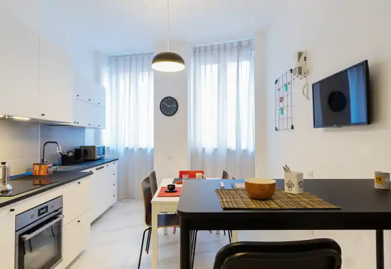 Bright modern kitchen with integrated appliances, a central dining counter and a small table set by large windows. Clean white cabinetry, black countertops and warm decorative accents create a welcoming space for cooking and casual dining.