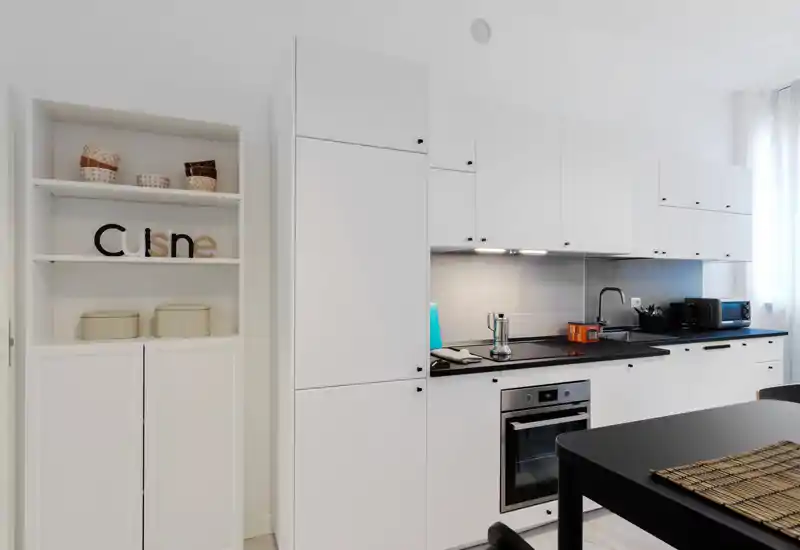 Modern white kitchen with integrated cabinets, built-in oven, black countertop and a small dining table in the foreground. Well-lit, tidy and styled with small accessories.