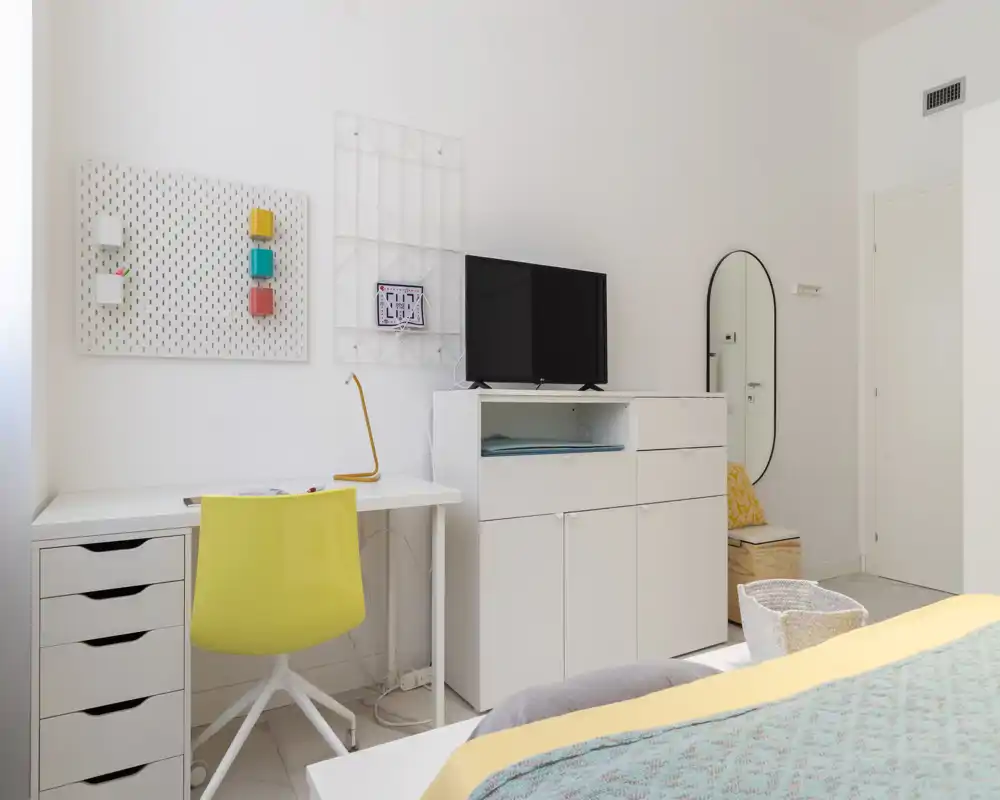 Bright modern bedroom with a visible bed covered in pastel textiles, a white desk with a yellow chair, and a TV on a low dresser — well lit and simply styled.