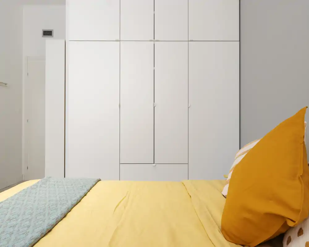 Bright, modern bedroom with a neatly made bed in the foreground and large white built-in wardrobes along the wall. Warm yellow bedding and cushions add a cozy accent to the minimalist space.