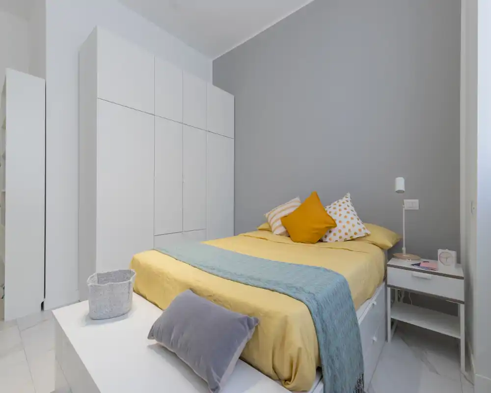 Bright, modern bedroom with a neatly made double bed, colorful cushions and a large white wardrobe. Soft natural lighting and minimalist décor create a fresh, welcoming atmosphere ideal for rental photos.