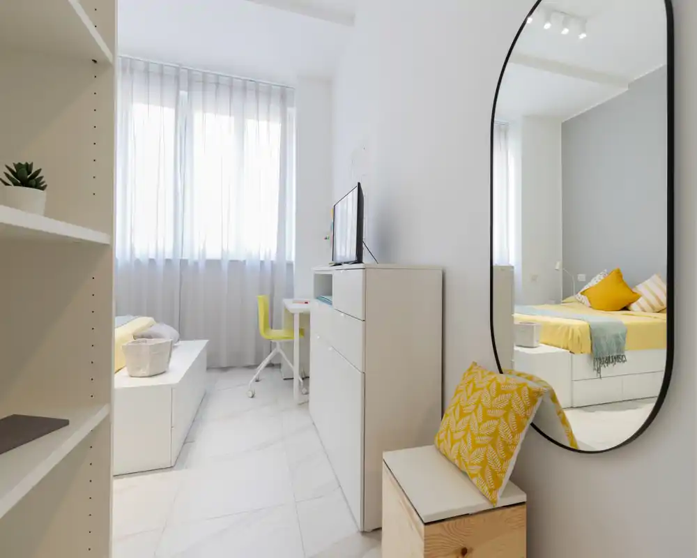 Bright, modern bedroom with a neatly made bed, soft yellow accents and a large mirror that reflects the bed and window light. The room features clean white furniture, a desk with a chair and pleasant natural lighting.