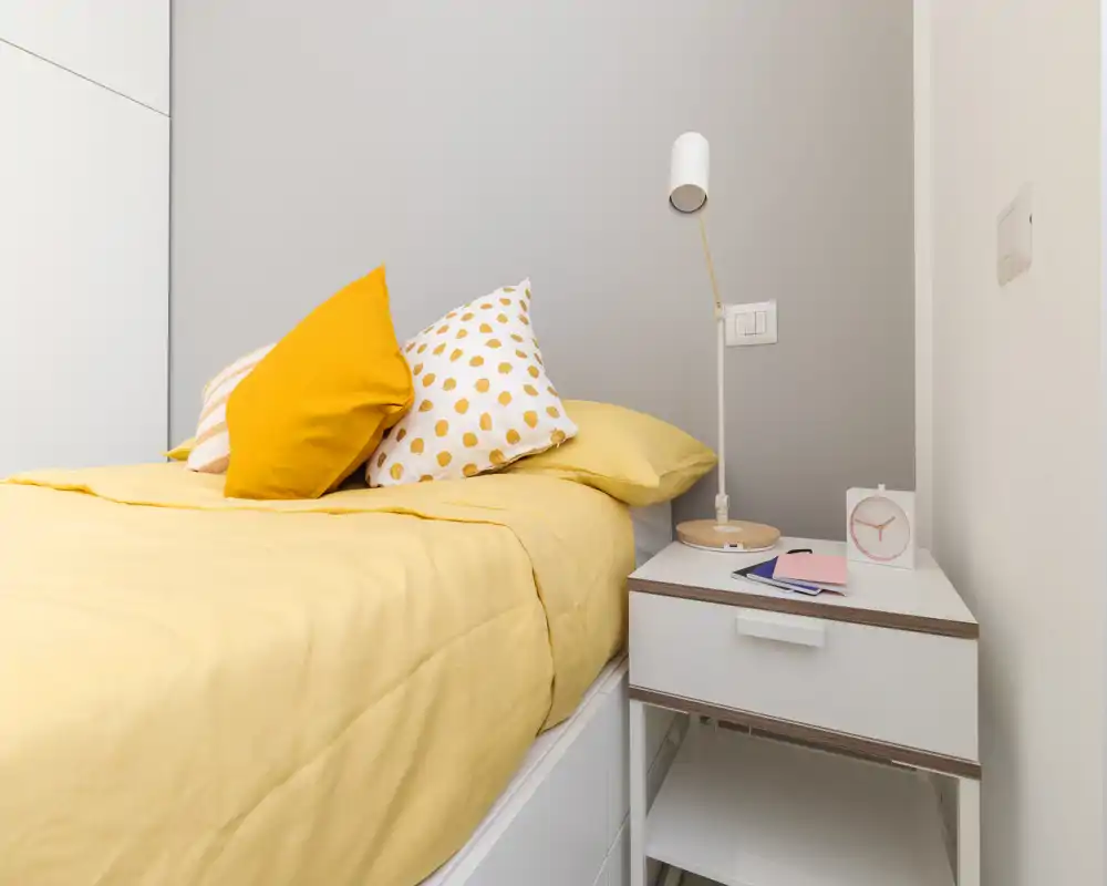 Bright, neatly staged bedroom corner with a yellow duvet and patterned cushions on a single bed beside a white nightstand and lamp — clean, modern and welcoming.