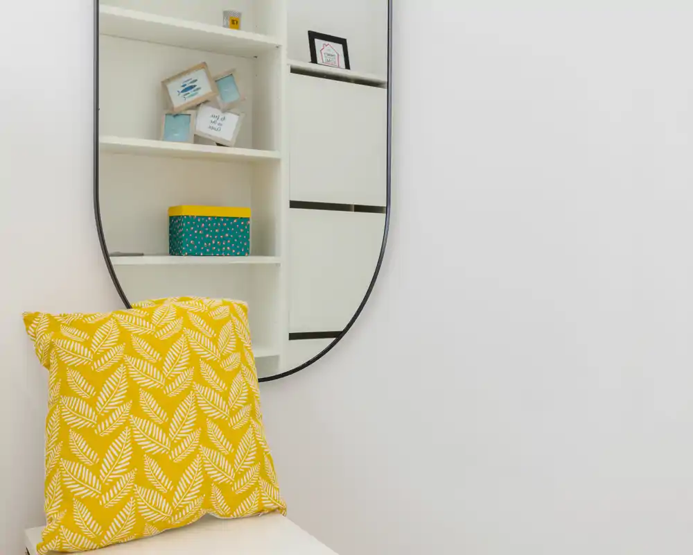 A close-up decorative corner showing a yellow patterned cushion on a small seat and a wall-mounted oval mirror reflecting shelving. The image focuses on decor details rather than a full room.