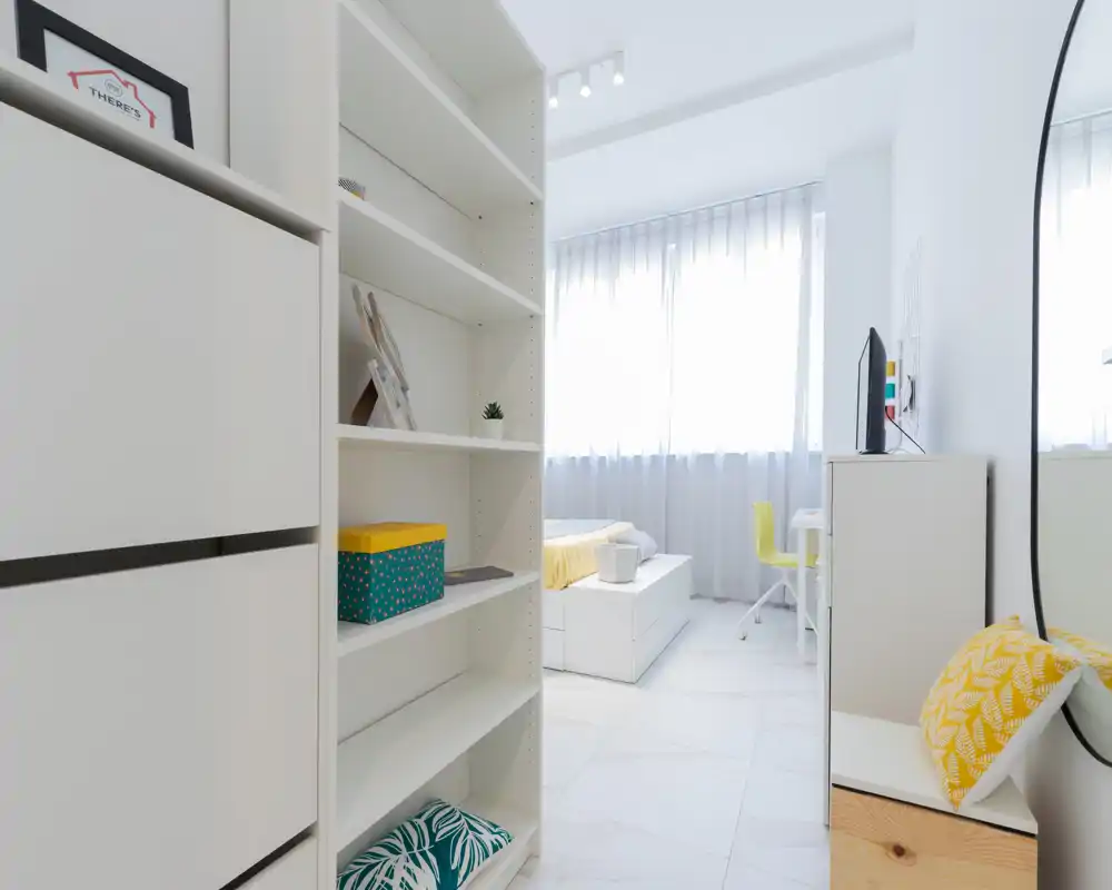Bright, minimalist bedroom with a neatly made single bed, white shelving and a small desk near the window — ideal for a student or single occupant.