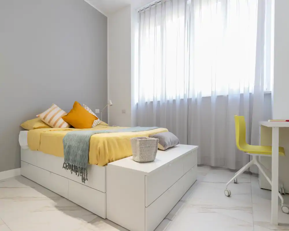 Bright, modern bedroom with a low platform bed, cozy throws and pillows, and a light-filled window with sheer curtains.