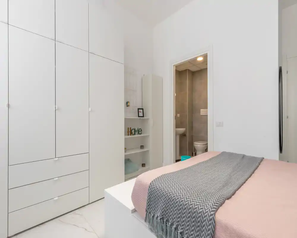 Bright, minimalist bedroom with a neatly made bed in the foreground and built-in white wardrobes along the wall. The ensuite bathroom is visible through a door, and soft textiles add a calm, modern feel.