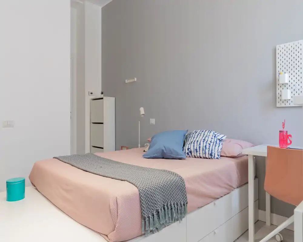 Modern, softly lit bedroom with a neatly made double bed, decorative pillows and a knitted throw. Clean, minimalist styling with a small desk and storage, ideal for rental listings that highlight comfort and contemporary design.